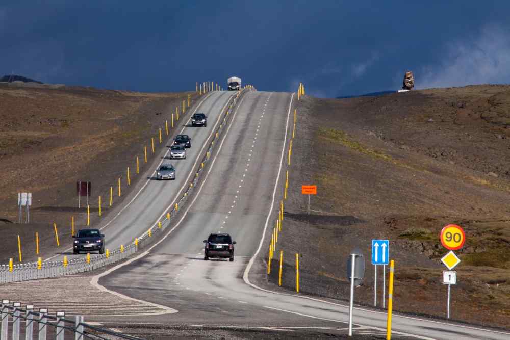 Auto che percorrono la Route 1 islandese accanto a un cartello con limite di velocità di 90 km/h in un remoto paesaggio vulcanico.