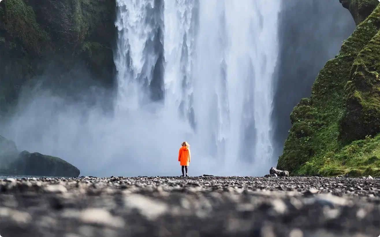 Viaggiatore con giacca arancione davanti a Skógafoss, simbolo della nostra missione di offrire esperienze memorabili.